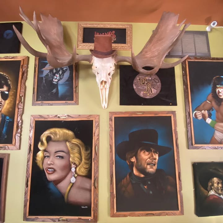 A wall of framed velvet paintings beside a moose skull wearing a top hat
